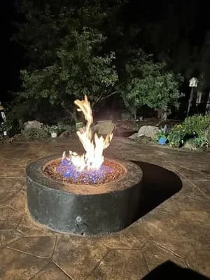 Circular stone fire pit with purple glass media and flame at night on stamped concrete patio